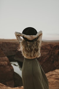 a woman wearing a hat and long dress standing on a cliff overlooking a canyon